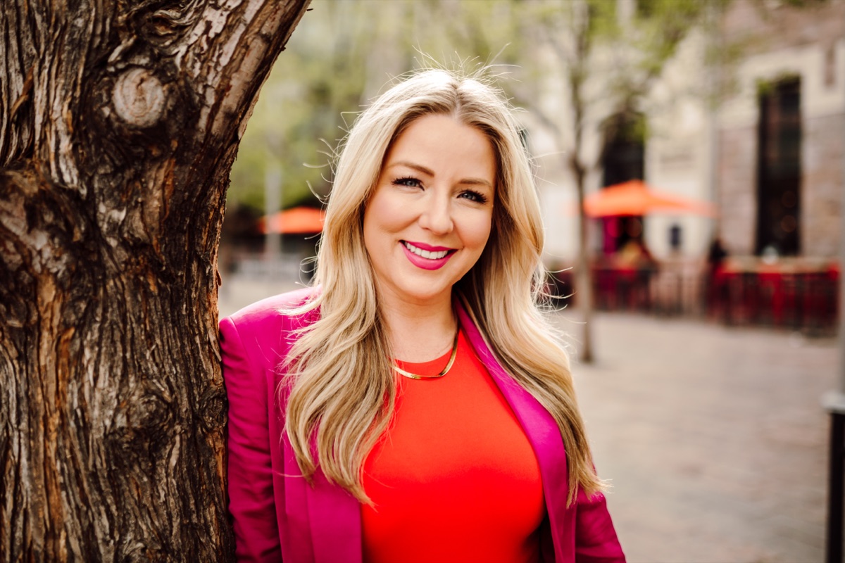 Headshot client in a magenta blazer over an orange top, leaning against a tree in a downtown plaza