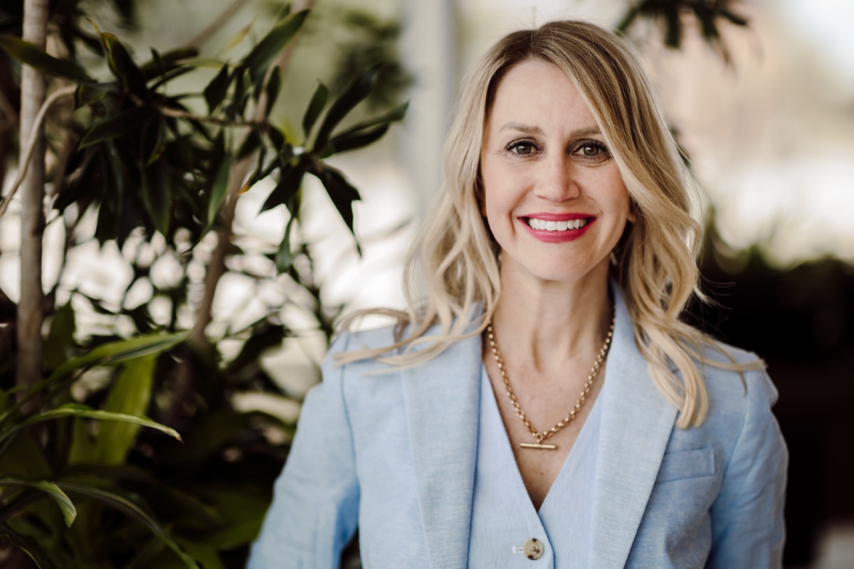 Headshot client in a light blue blazer and gold necklace, smiling among leafy plants