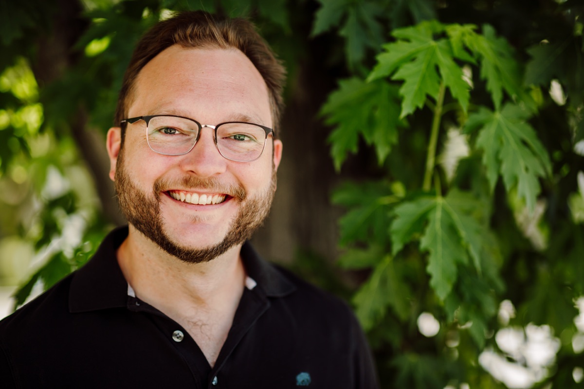 Headshot client in glasses and a black polo, smiling in front of sunlit green leaves