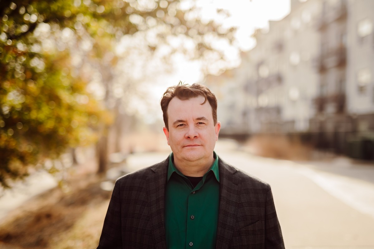 The same client in a green shirt and plaid blazer on a tree-lined path