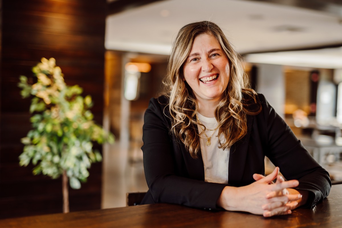Client in a black blazer over a white tee with gold necklaces, laughing at a wooden table