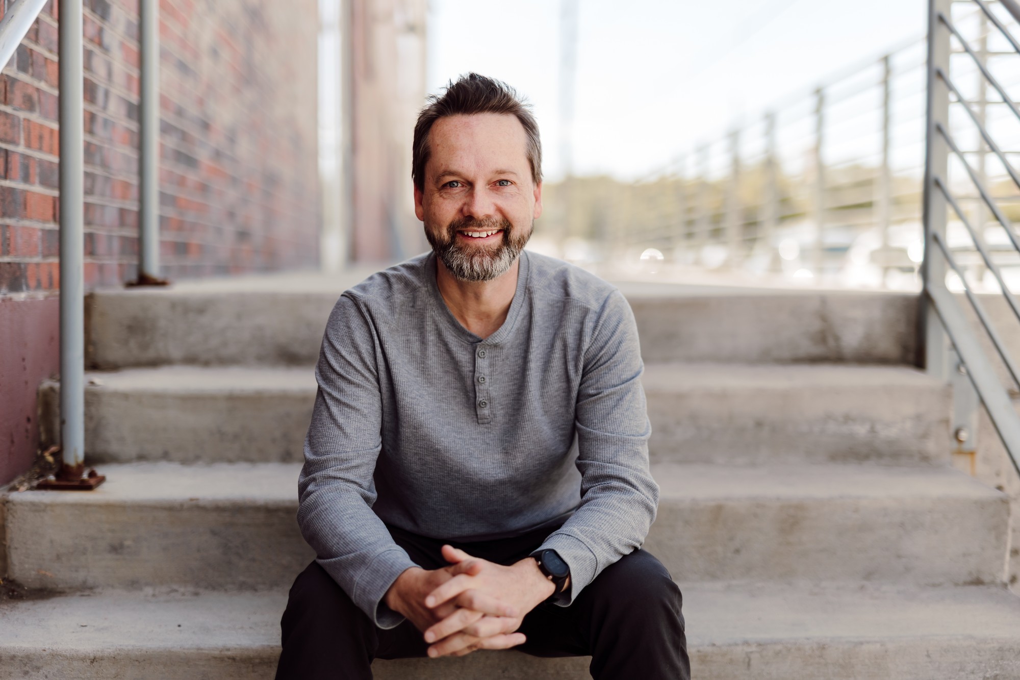 Headshot client with a grey beard in a grey henley, seated on concrete steps by a brick wall