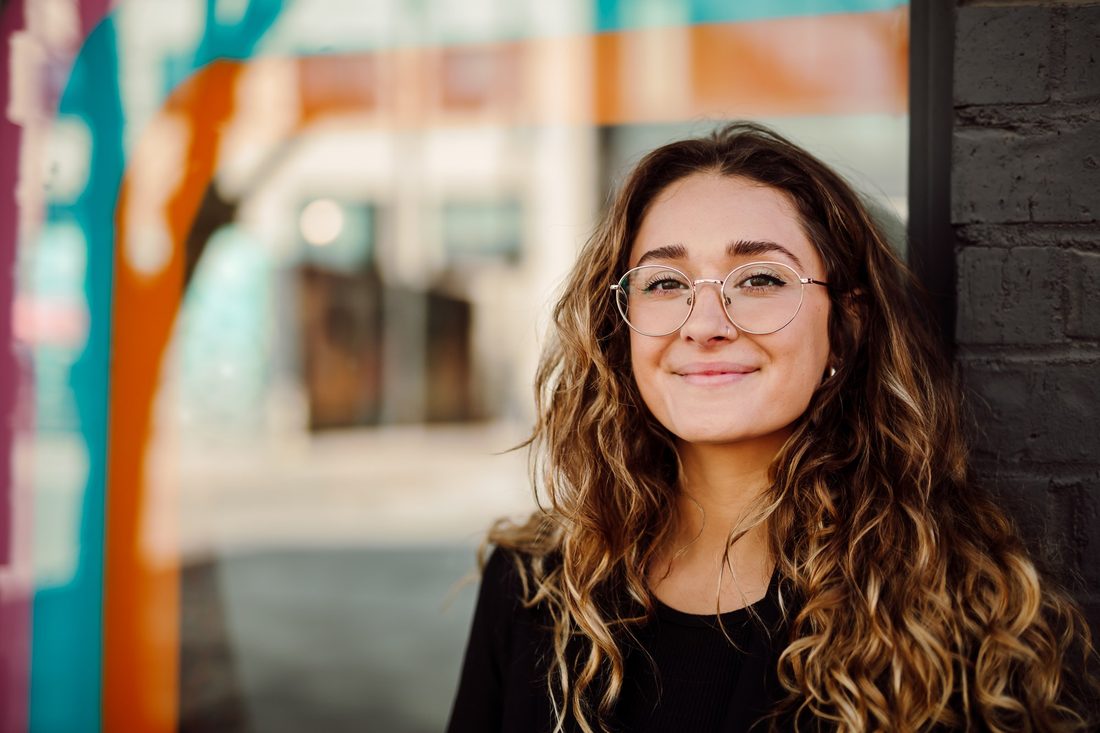 Headshot client in glasses and a black top, smiling in front of a teal and orange painted wall